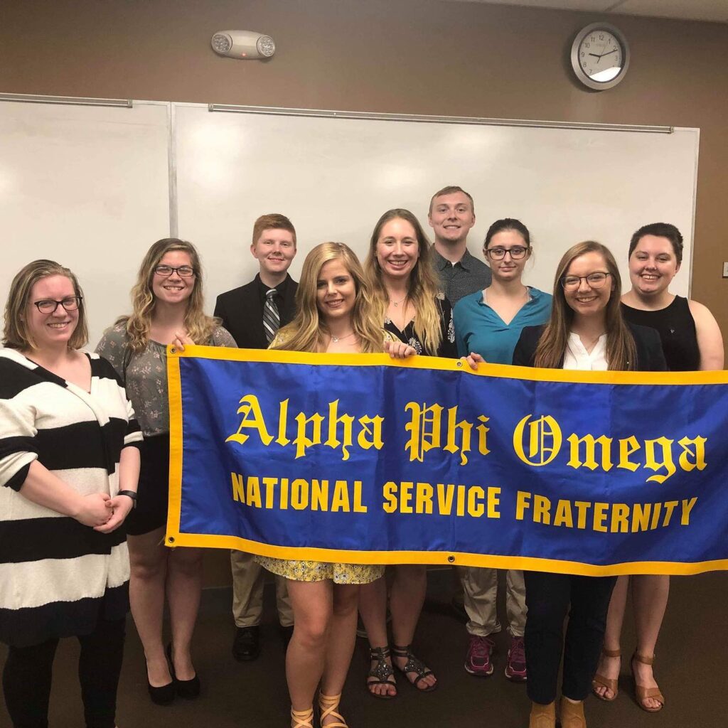 The Alpha Eta Lambda chapter of Alpha Phi Omega at Eureka College poses as a group following a ceremony in Fall 2019. The members in the front row hold a blue banner with a gold border and lettering that reads "Alpha Phi Omega National Service Fraternity"