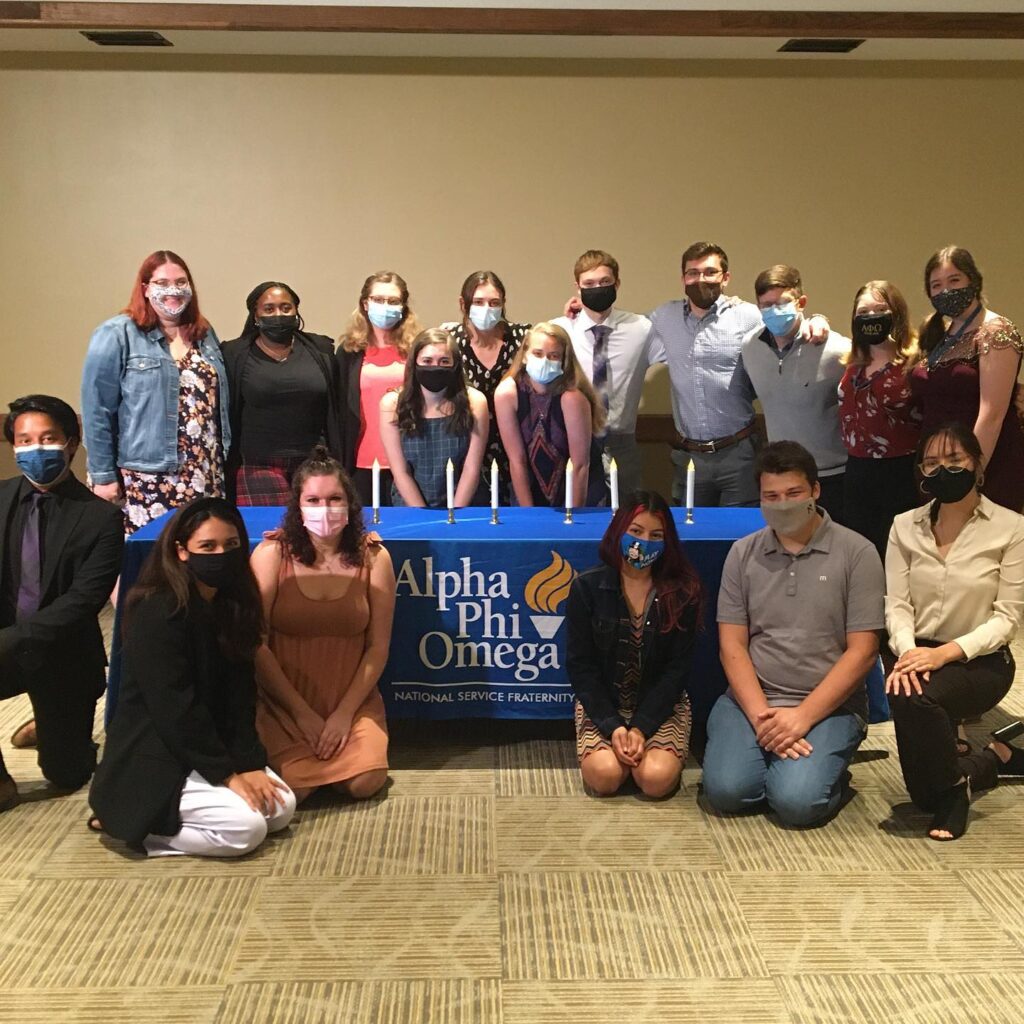 The Theta Epsilon chapter of Alpha Phi Omega at Illinois State University poses as a group following a ceremony in Fall 2021. They are grouped around a table with a blue cloth and candles with a banner that reads "Alpha Phi Omega National Service Fraternity"