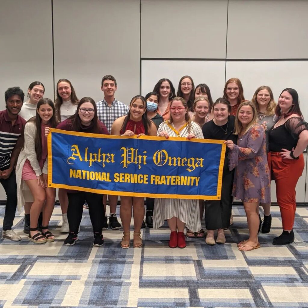 The Eta Epsilon chapter of Alpha Phi Omega at Millikin University poses as a group following a ceremony in Spring 2022. The members in the front row hold a blue banner with a gold border and lettering that reads "Alpha Phi Omega National Service Fraternity"