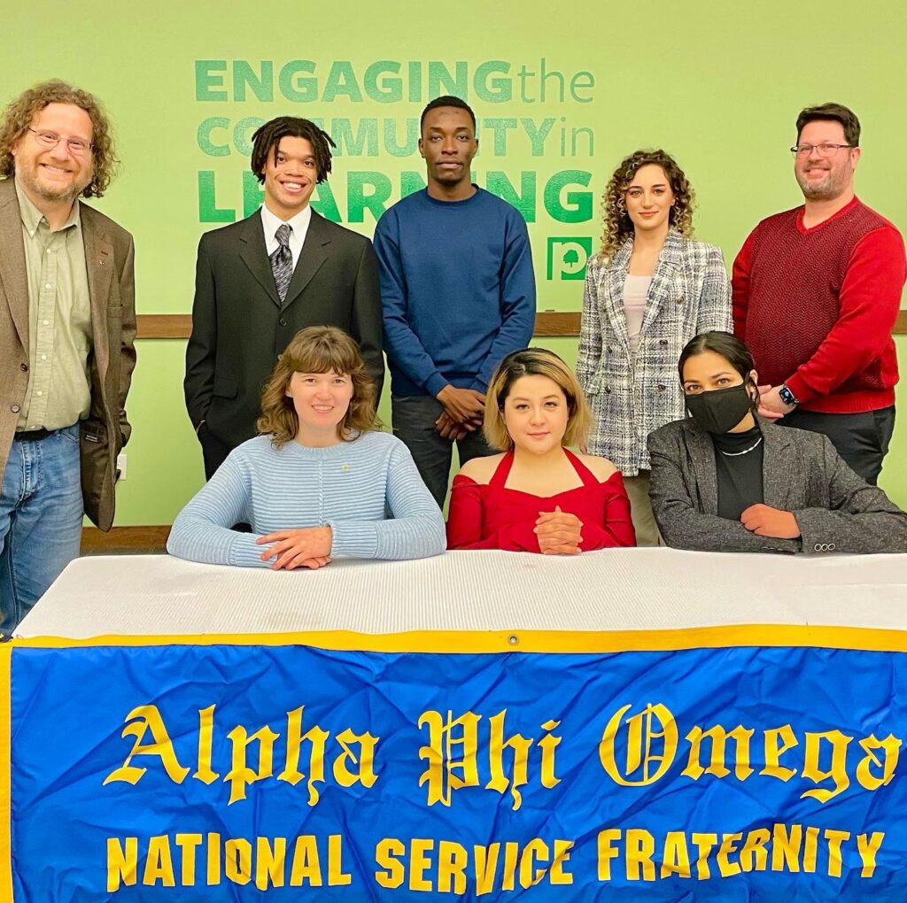 The Alpha Zeta Omega chapter of Alpha Phi Omega at Parkland College poses as a group following a ceremony in Spring 2022. The members are seated and standing behind a table that holds a blue banner with a gold border and lettering that reads "Alpha Phi Omega National Service Fraternity"